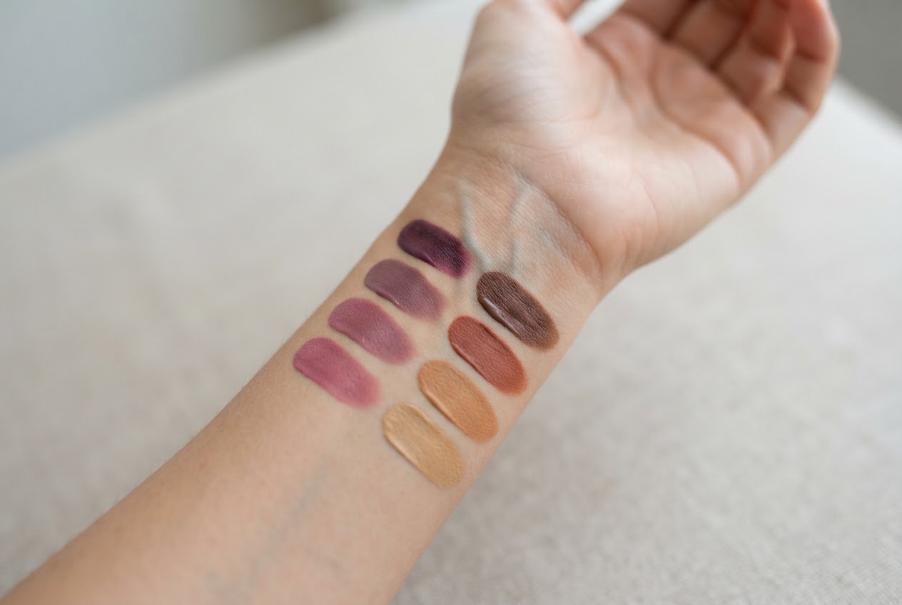 A hand held up in natural light, showing the inner wrist with visible veins, and several makeup swatches in cool and warm tones on the forearm.