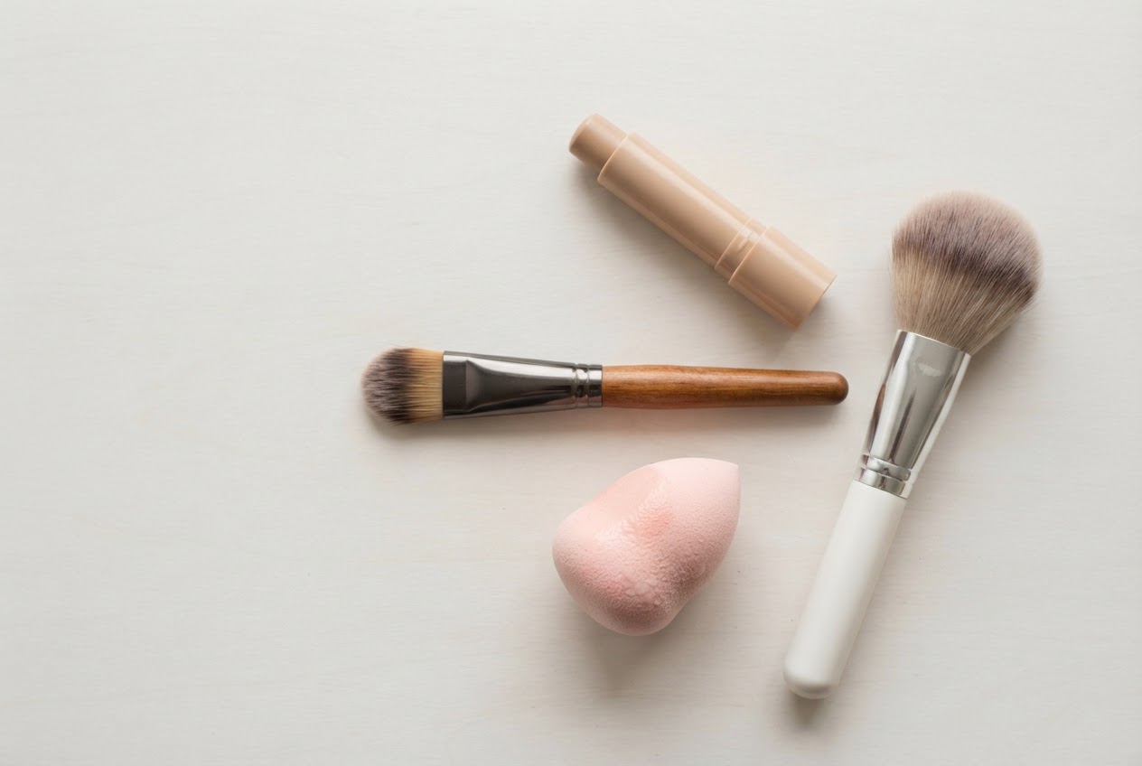 A flat lay of essential camouflage tools: flat synthetic brush, damp beauty blender, powder brush, and a camouflage stick.