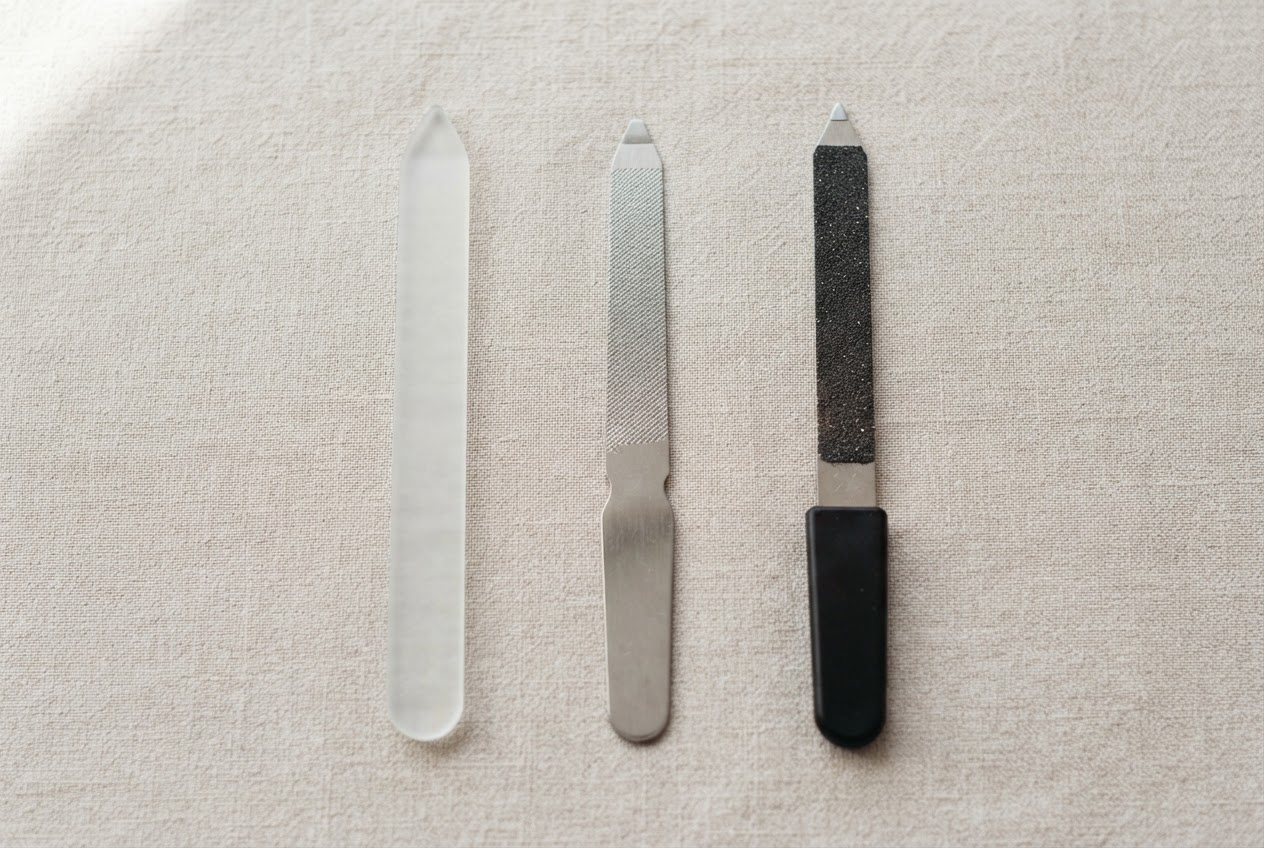 Drei verschiedene Nagelfeilen aus Glas, Metall und Saphir liegen nebeneinander auf einem hellen Tuch.