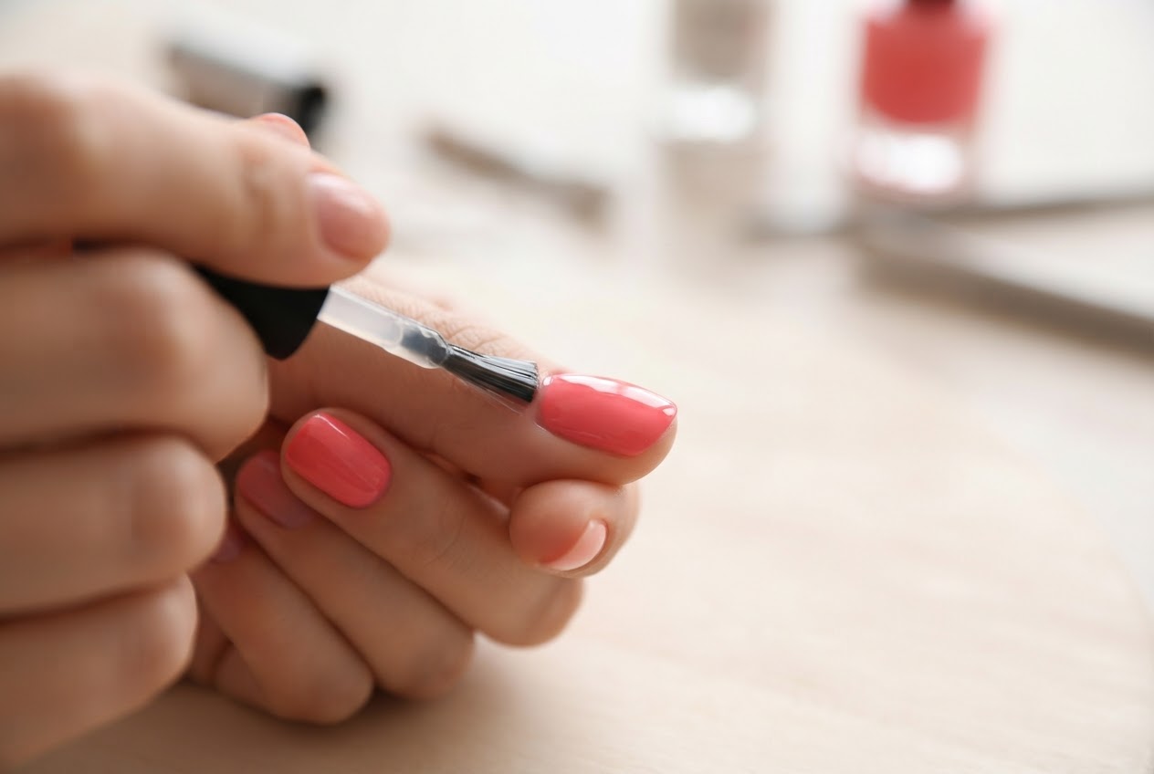 A hand applying a clear top coat with a brush to a painted nail, the brush leaving a glossy layer.
