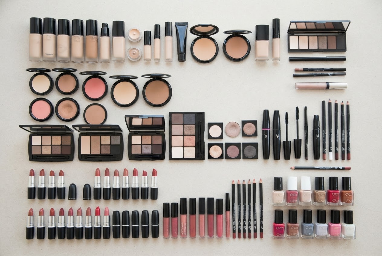 A flat lay of various MC Marie Christine makeup products including foundation, concealer, powder, eyeshadow, mascara, eyeliner, lipstick, lip gloss, lip liner, blush, bronzer, and nail polish on a clean surface.