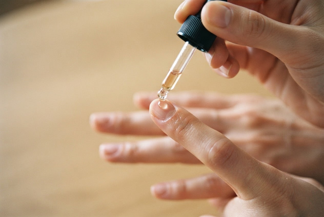 A hand applying a drop of nail oil from a bottle to a fingernail, with focus on the dropper and nail.