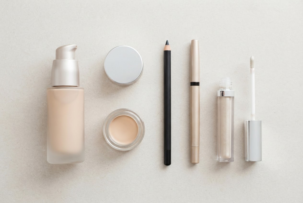 A flat lay of five unbranded make-up products: a foundation pump, a concealer jar, an eyeliner pencil, an eyeshadow pen, and a lip gloss tube on a light surface.
