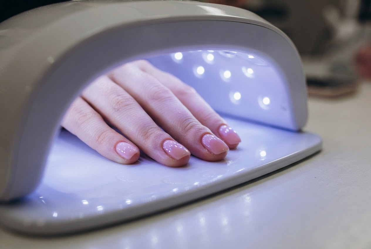 A close-up of fingers placed inside a UV nail lamp, the lamp emitting a blue-purple glow, skin around nails visible.