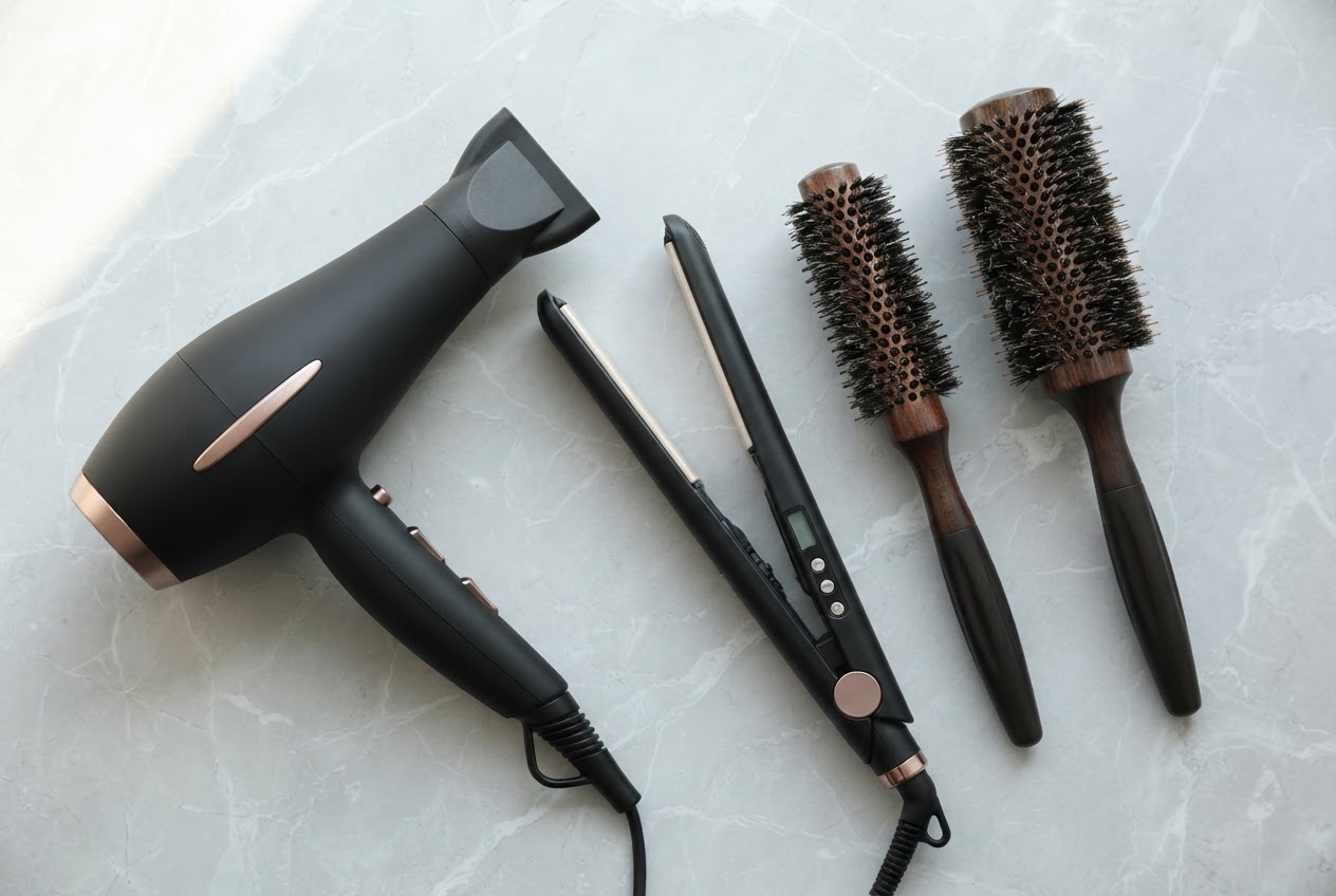 Flat lay of professional hair tools: an ion hair dryer, a flat iron with temperature control, and two round brushes of different sizes.