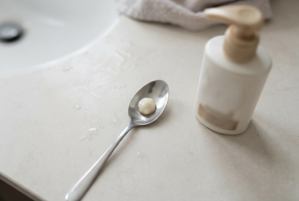 A half teaspoon of shampoo next to a full-sized bottle, showing a tiny portion.