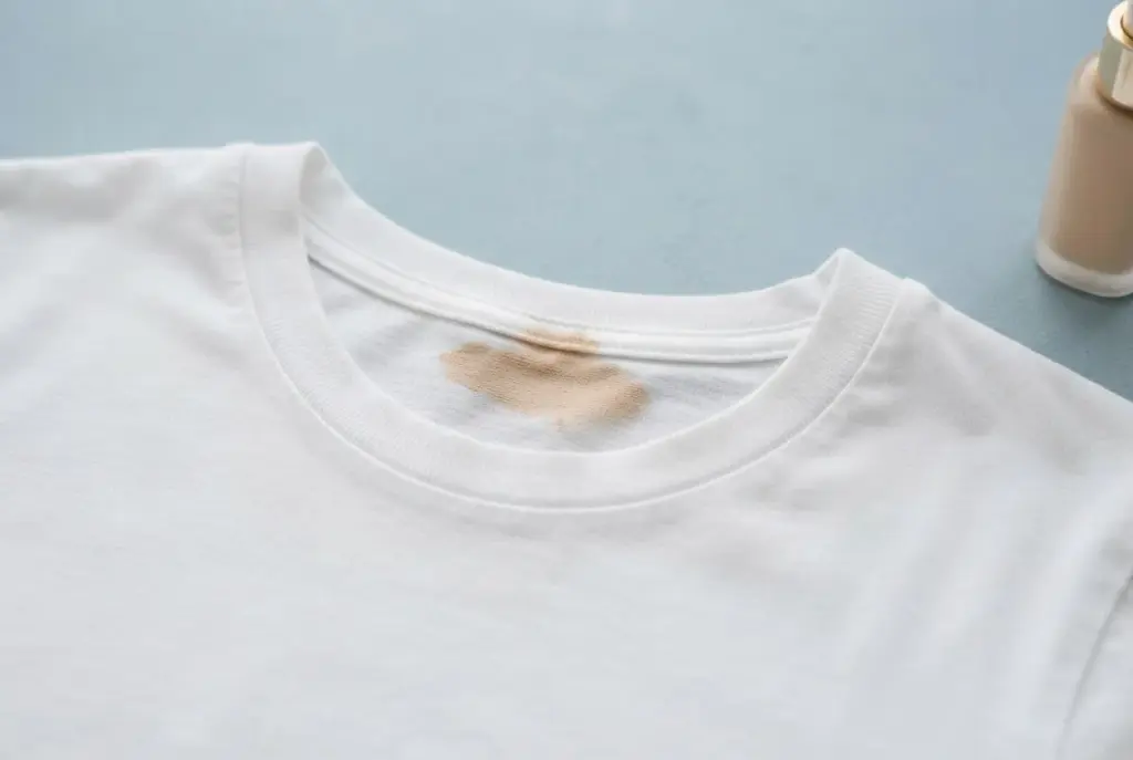 Ein beiger Make-up-Fleck am Ausschnitt eines weißen Baumwollshirts; daneben steht eine schlichte Foundationflasche.
