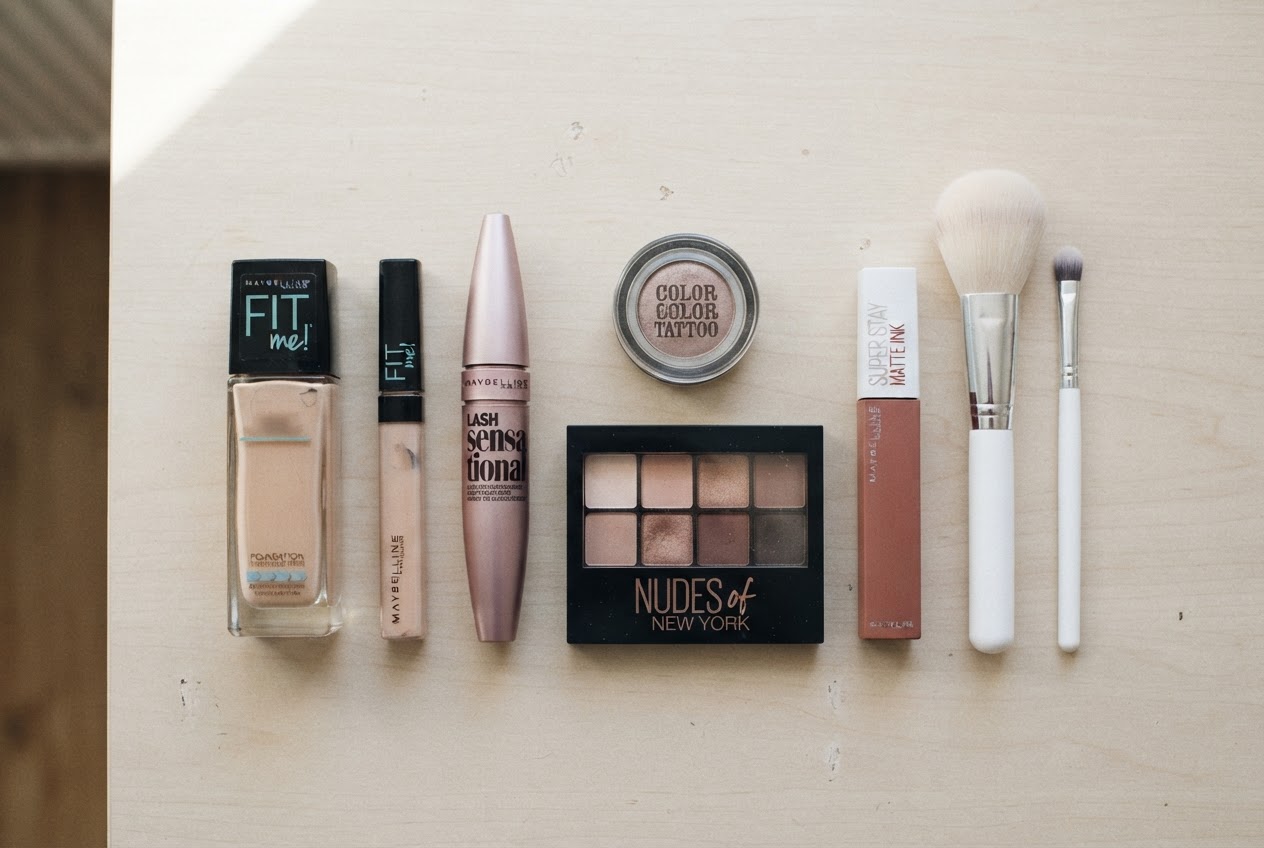 A flat lay of Maybelline makeup products: foundation, concealer, mascara, eyeshadow palette, lipstick, and brushes on a clean surface.