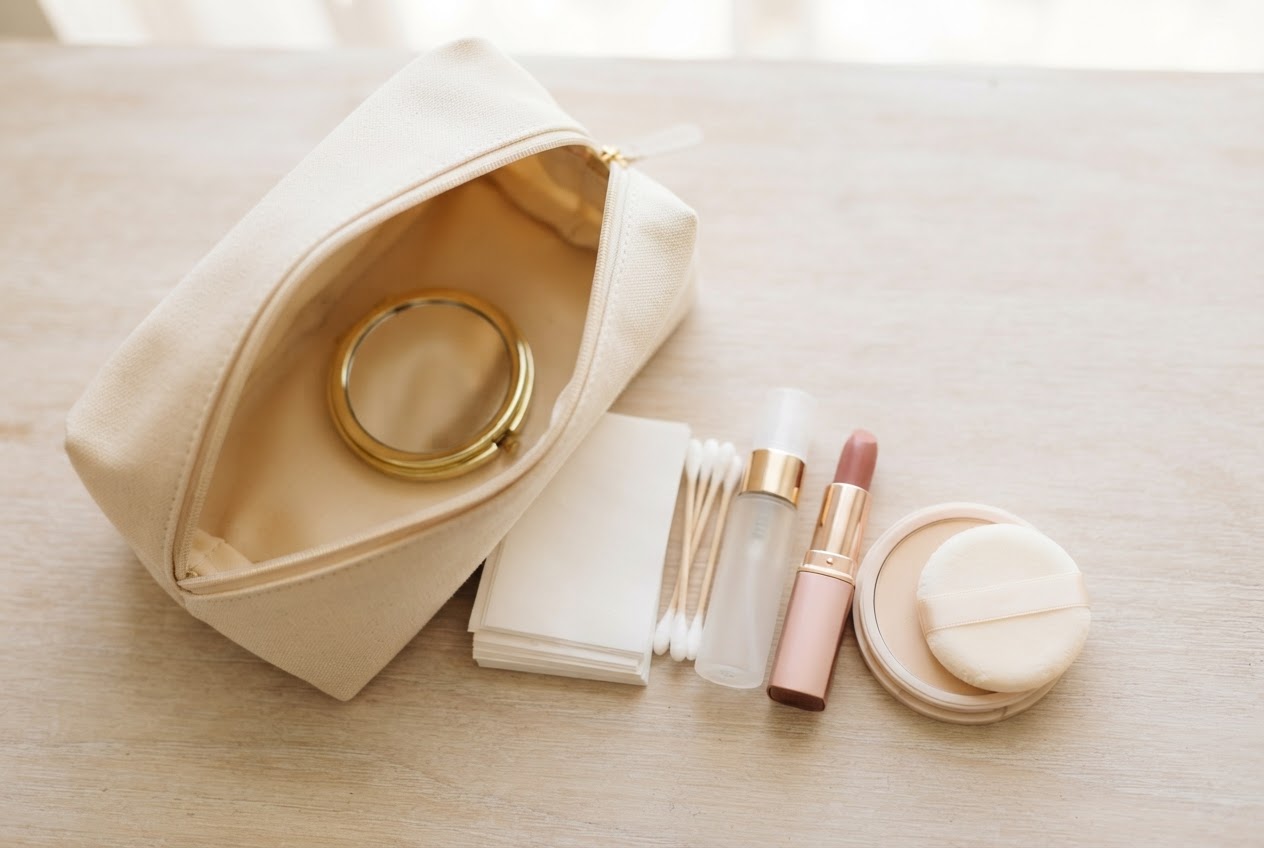 Ein offenes Make-up-Täschchen mit Wattestäbchen, Blotting-Papier, Kompaktpuder, Lippenstift, Mini-Setting-Spray und kleinem Spiegel.