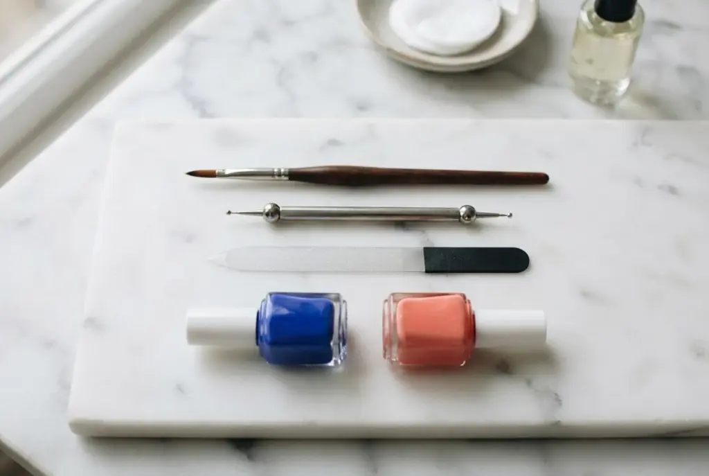 Ein professionell arrangiertes Set aus Nagelpinseln, einem Dotting-Tool, einer Nagelfeile und zwei Nagellacken in Blau und Koralle auf einer sauberen, hellen Oberfläche.
