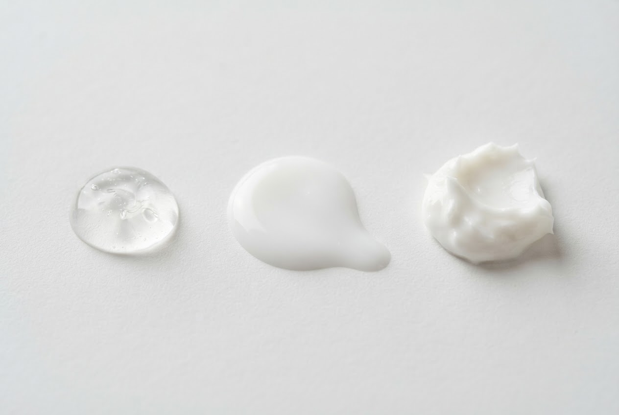 Three small dabs of skincare products on a white surface: a clear gel, a milky fluid, and a thick cream, showing different consistencies.