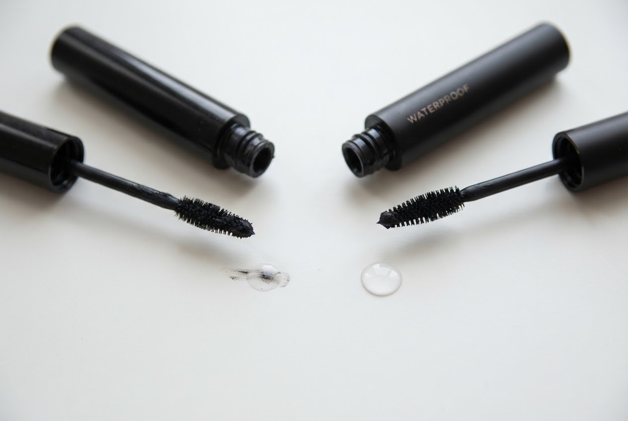 Zwei Mascara-Tuben liegen nebeneinander, eine mit wasserfester Formel, die andere mit normaler, davor ein Tropfen Wasser zur Verdeutlichung.